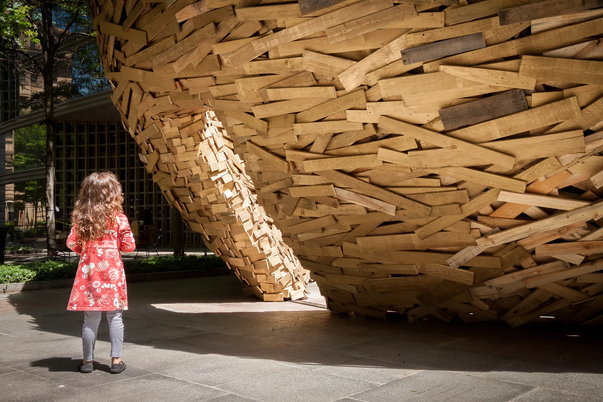 A child in a red coat looking up at a large sculpture of stacked wooden slats in a sunlit plaza with trees.